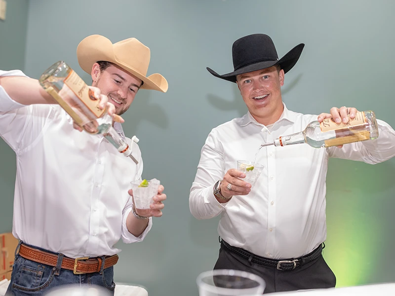 Two men in cowboy hats pour beverages into cups.