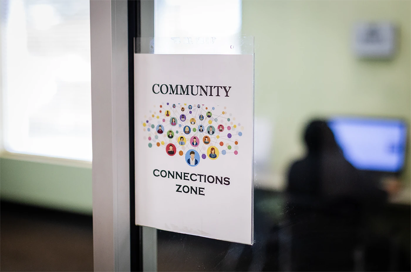 Sign reading "Community Connections Zone" taped to the window of an interior office space.